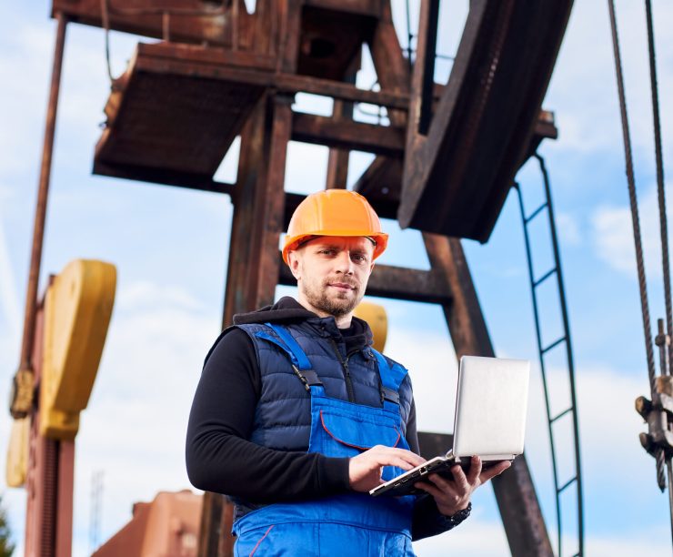 Vertical portrait of oil man, wearing blue overalls and orange helmet, with a laptop, standing with his back to an oil rig, checking oil pumping unit, making notes in his computer